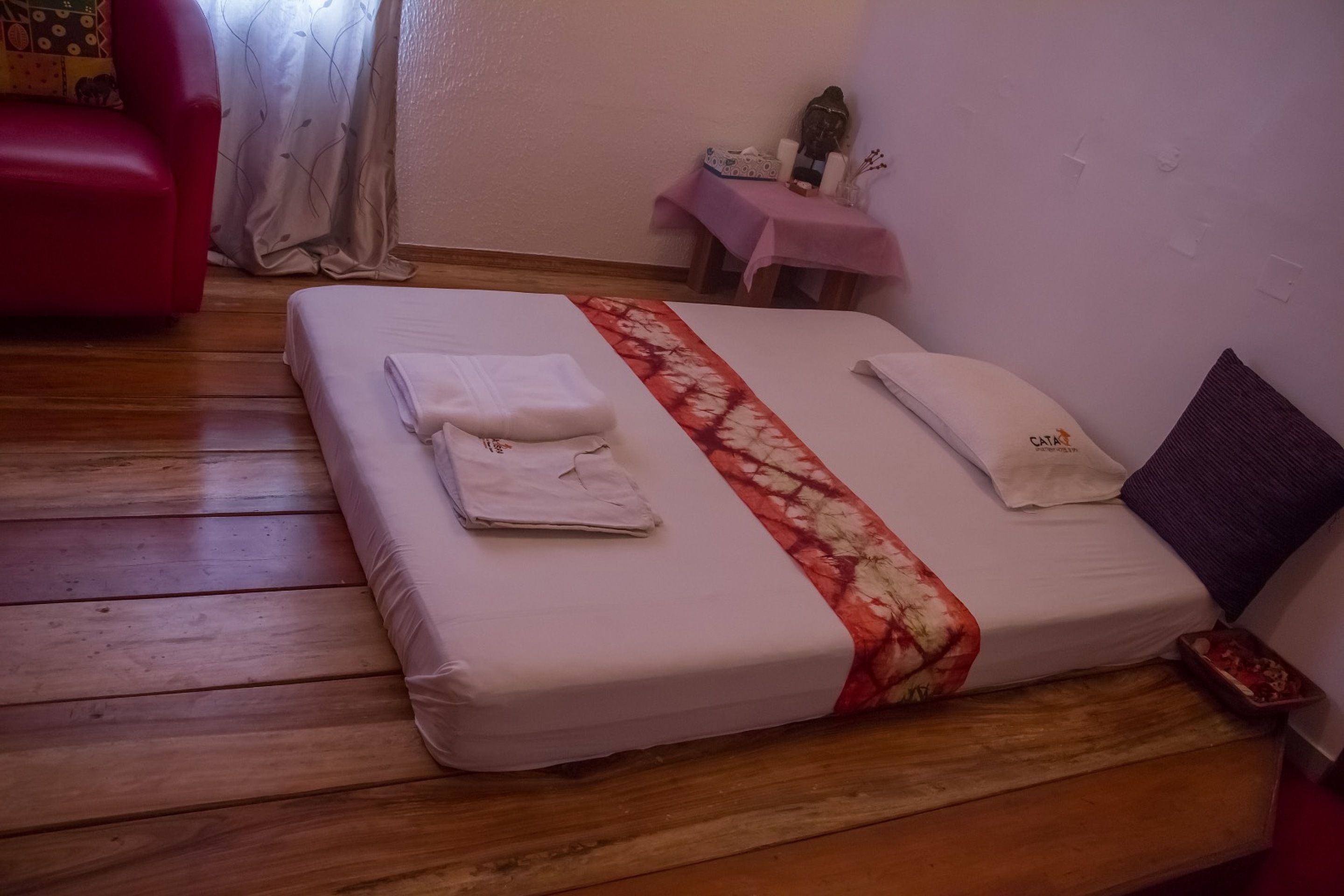 A neatly made bed with a floral bedspread, a red chair, and a small table with a pink cloth.