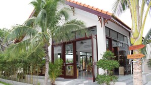 Un edificio moderno con fachada de vidrio, un camino de piedra que lleva a la entrada, y un cartel que indica un restaurante y una playa cerca.
