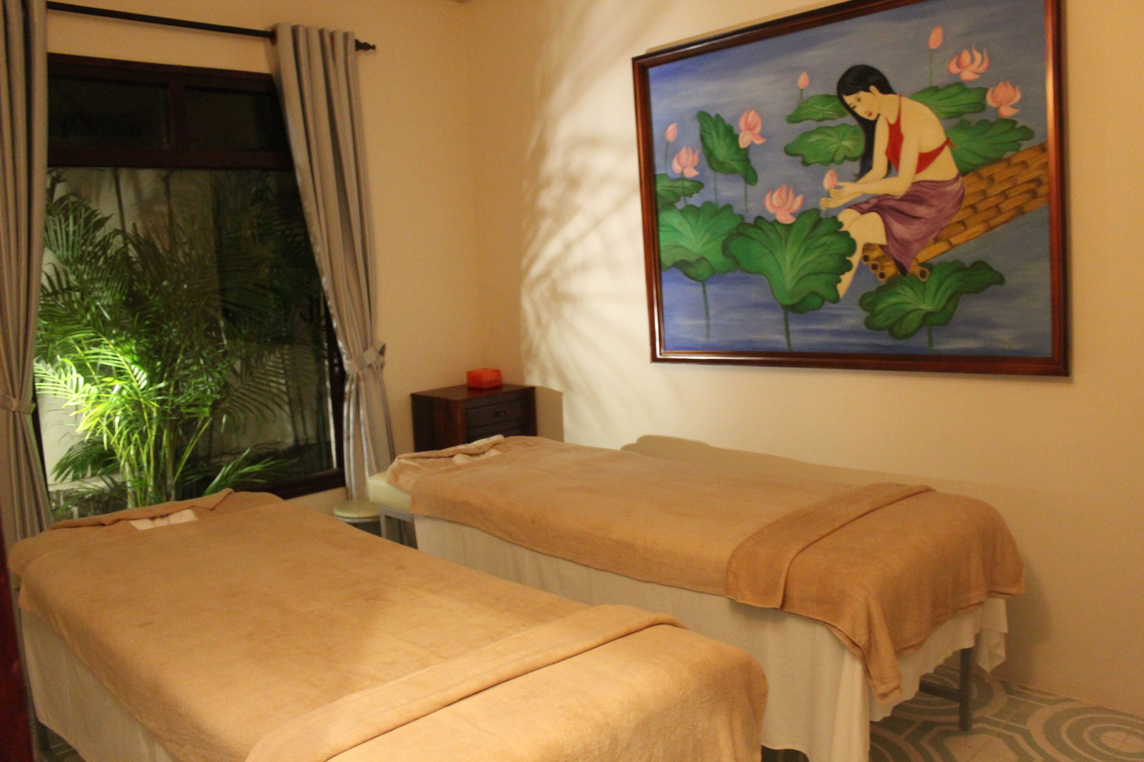 A room with two massage tables, a painting of a woman with lotus flowers, a window with curtains, and a small table with a red object.