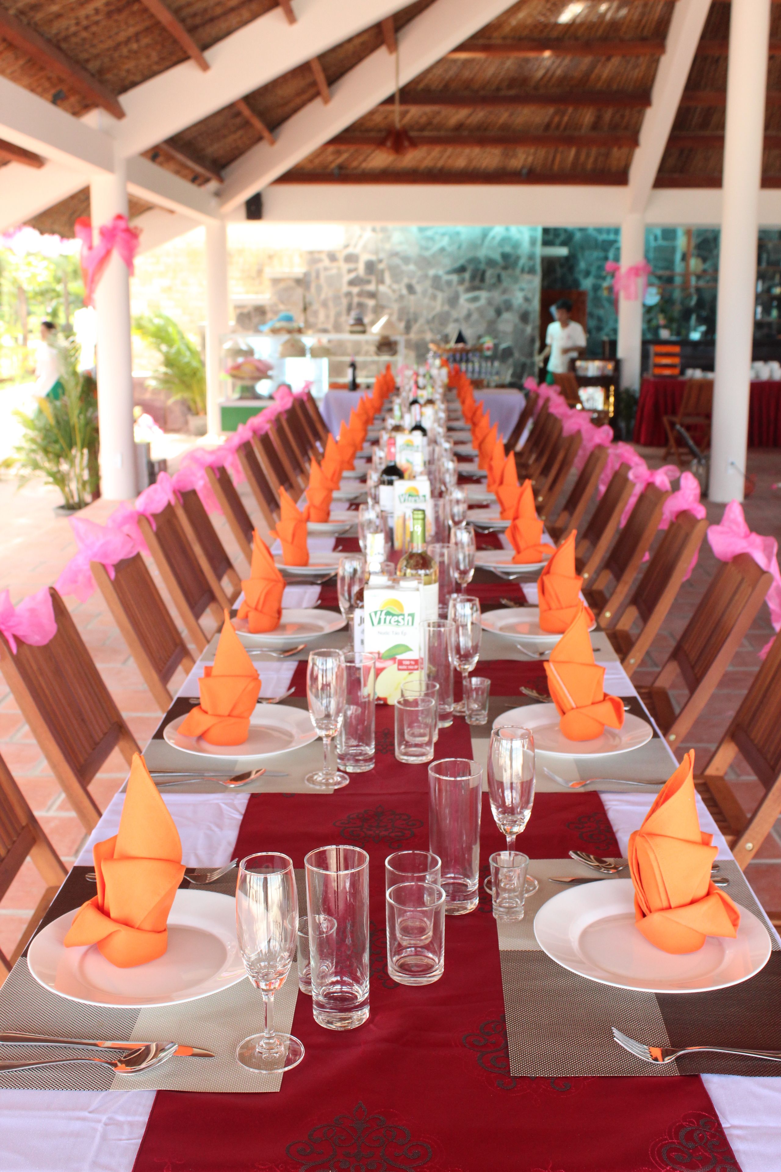 Una larga mesa de comedor dispuesta con servilletas anaranjadas, copas de cristal transparentes y platos blancos.