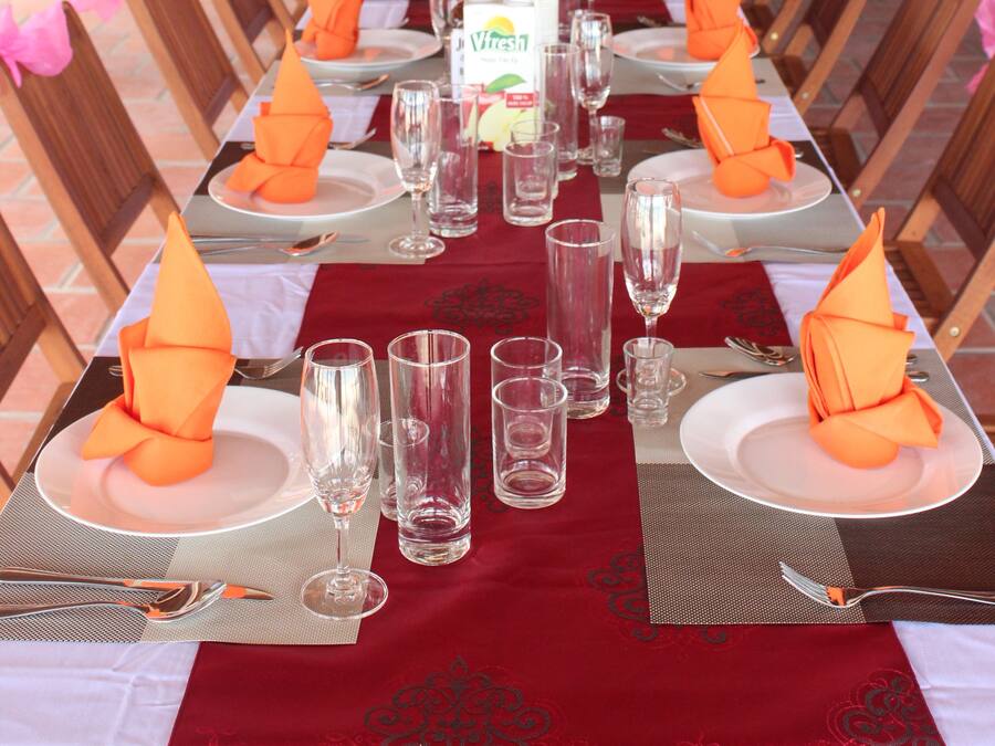 Una larga mesa de comedor dispuesta con servilletas anaranjadas, copas de cristal transparentes y platos blancos.