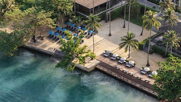 An aerial view of a resort with a beach area, lounge chairs, umbrellas, and palm trees.