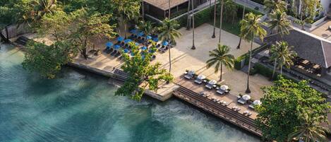 An aerial view of a resort with a beach area, lounge chairs, umbrellas, and palm trees.