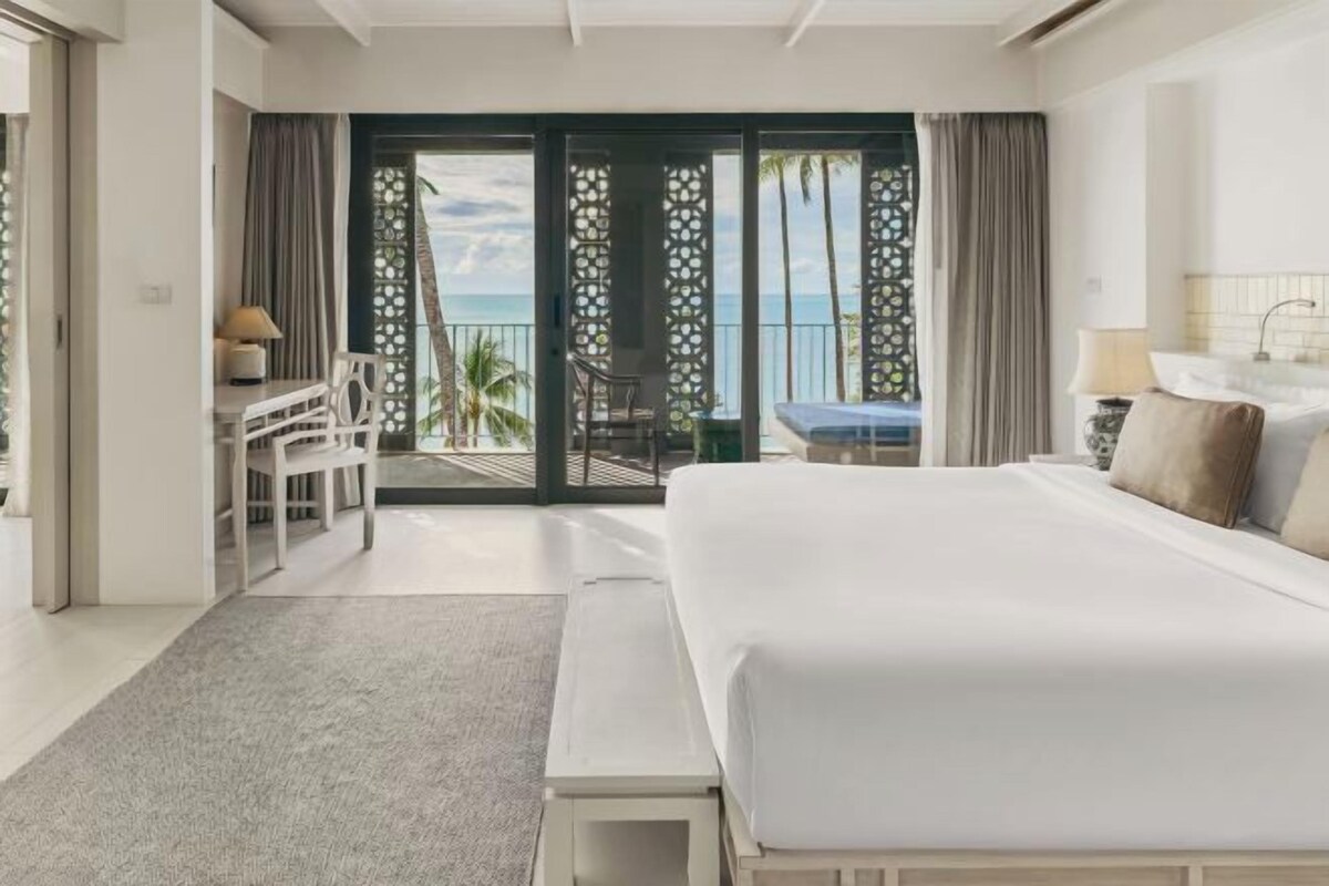 A hotel room with a large bed, a desk, a chair, and a view of the ocean through a sliding door.