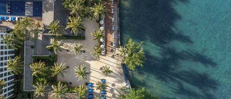Vista aérea de un hotel frente a la playa con un patio arenoso, palmeras y una zona de piscina.