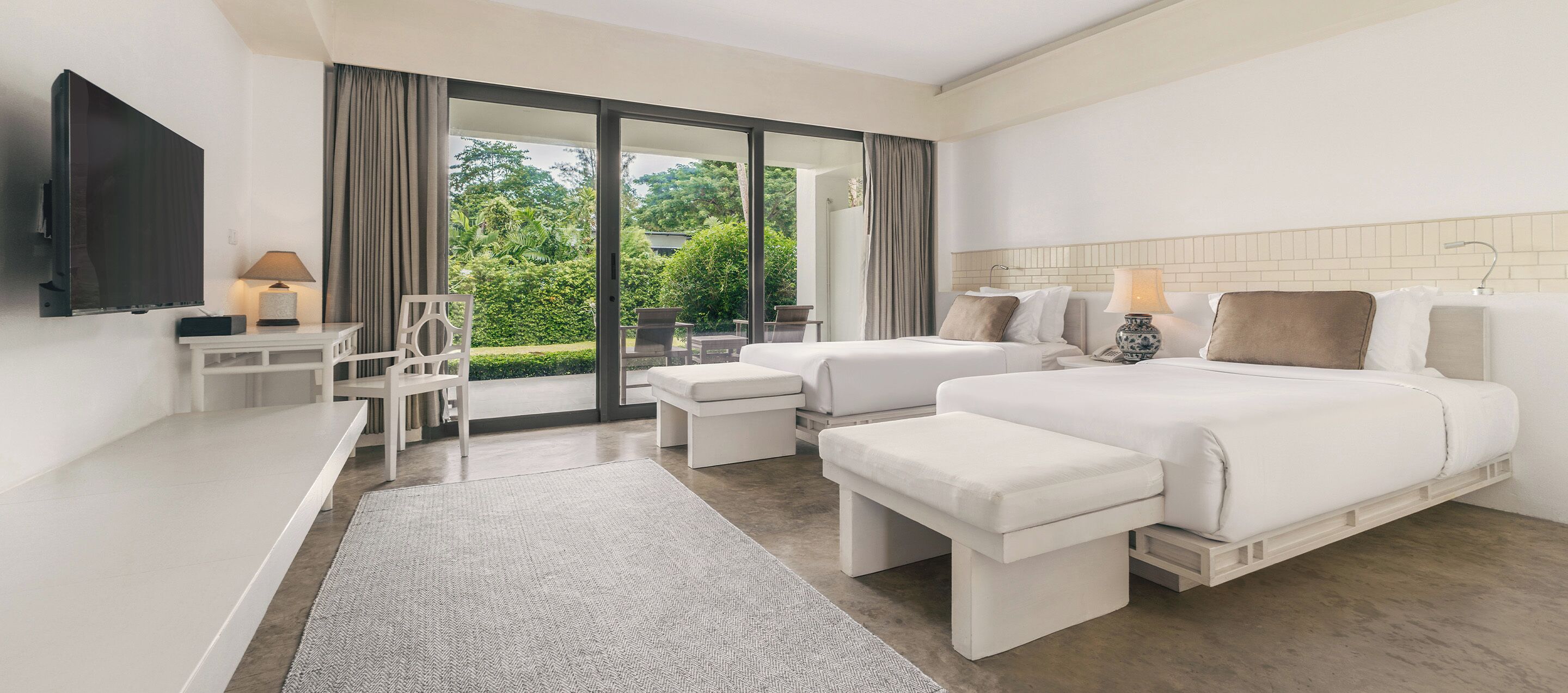 A hotel room with a large bed, a smaller bed, a desk with a TV, and a balcony with a view of greenery.