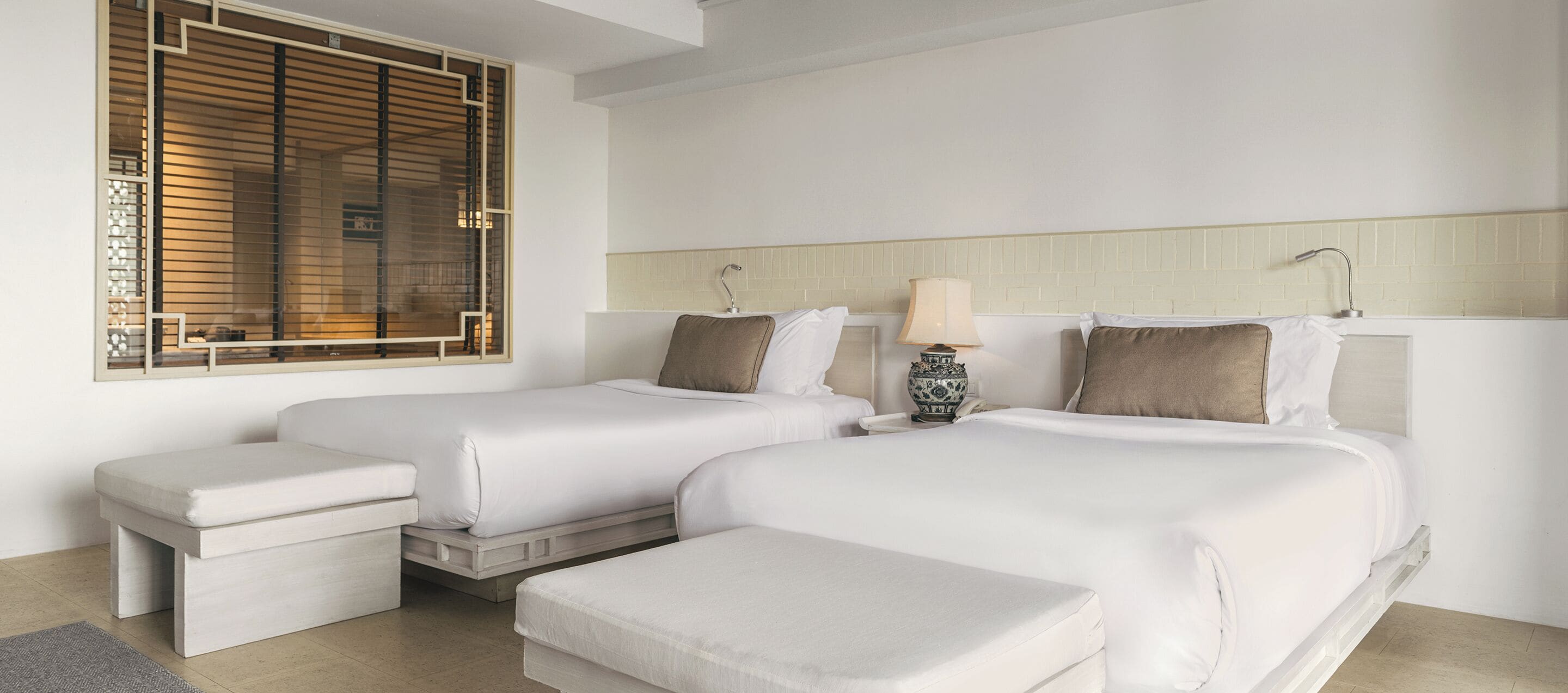 A hotel room with two beds, a small table, a bench, a window with blinds, and a decorative vase on the nightstand.