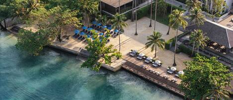 An aerial view of a resort with a beach area, lounge chairs, umbrellas, and palm trees.