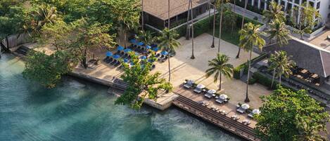 An aerial view of a resort with a beach area, lounge chairs, umbrellas, and palm trees.