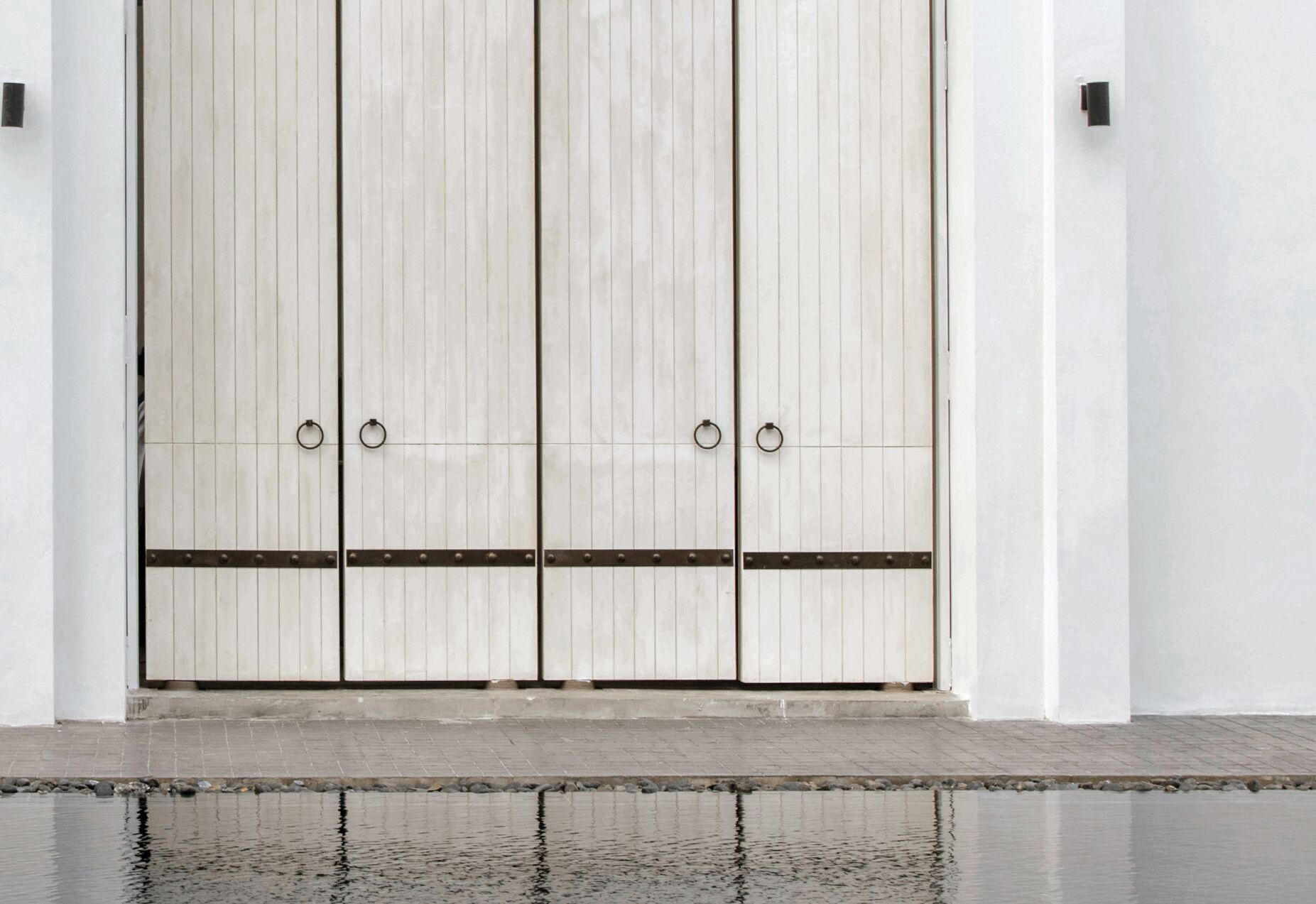 A large, white, double door with metal rings and a horizontal bar, set against a plain wall with a wet ground in front.