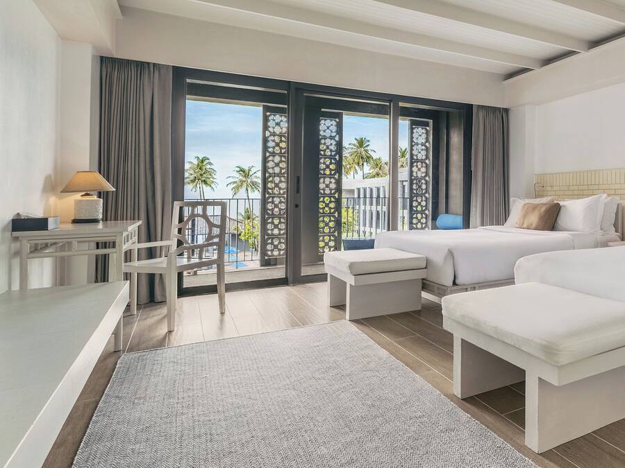 Habitación de hotel con dos camas, un escritorio, una silla, un balcón con vistas a palmeras y un ventanal grande.