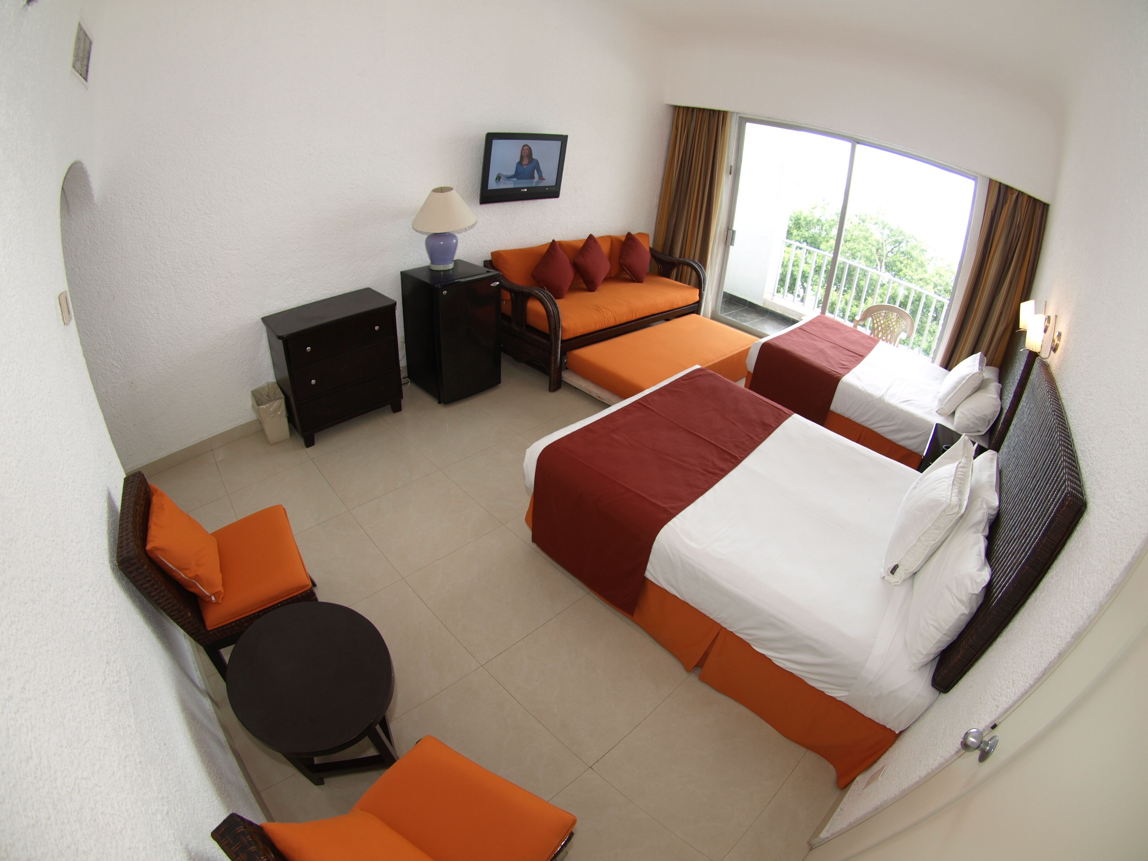 Une chambre d’hôtel avec deux lits, un canapé, une petite table et une chaise. Il y a un téléviseur accroché au mur et une fenêtre avec des rideaux.