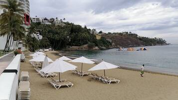 A beach with white umbrellas and lounge chairs, a sandy shore, and a building with balconies overlooking the sea.