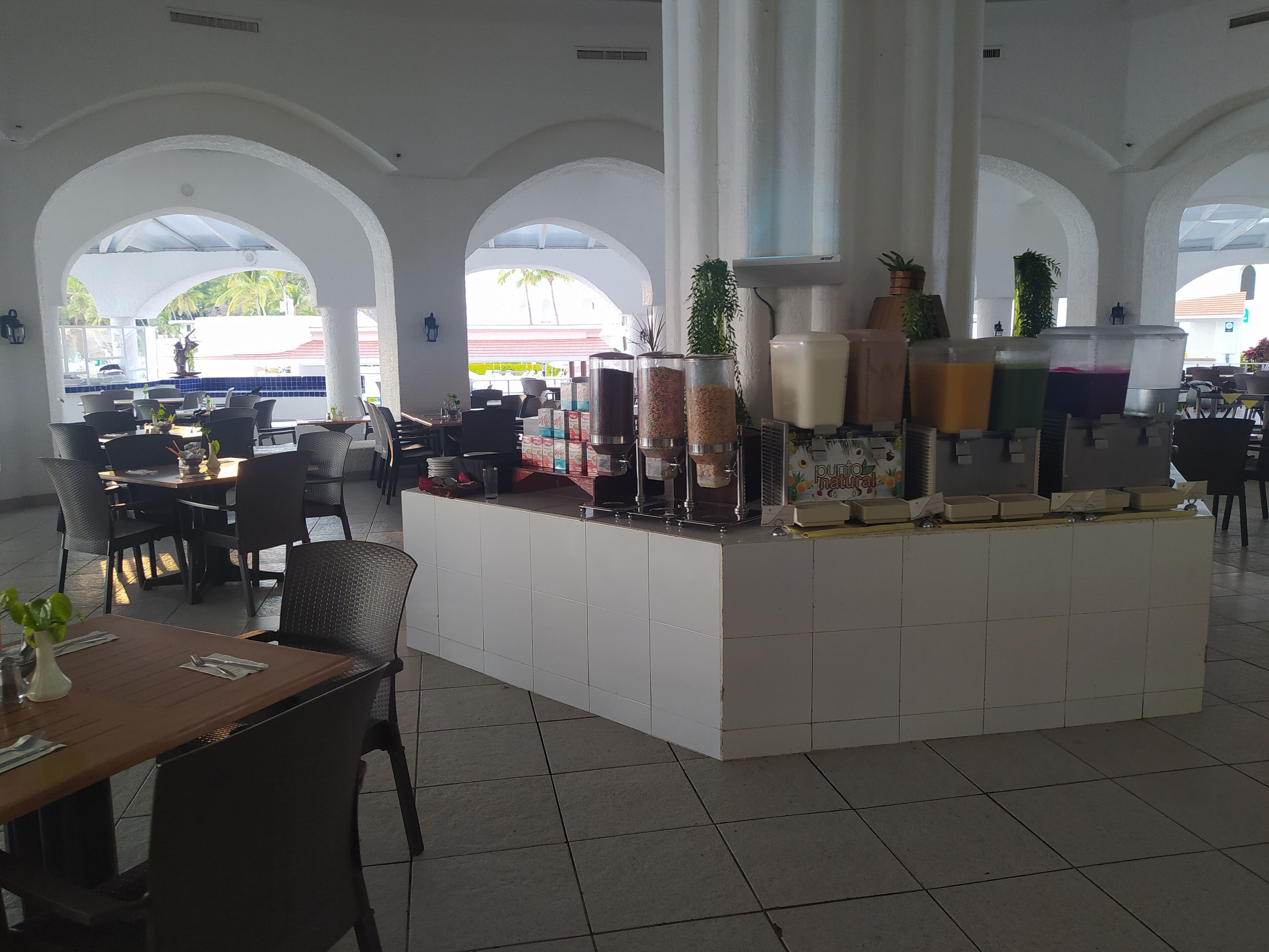 Un espacio de cafetería amplio y luminoso, con una barra central, diversas opciones de asientos y grandes ventanales.