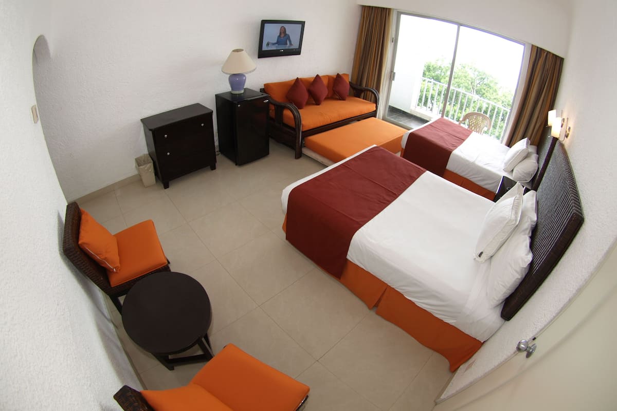 A hotel room with two beds, a sofa, a small table, and a chair. There is a TV on the wall and a window with curtains.