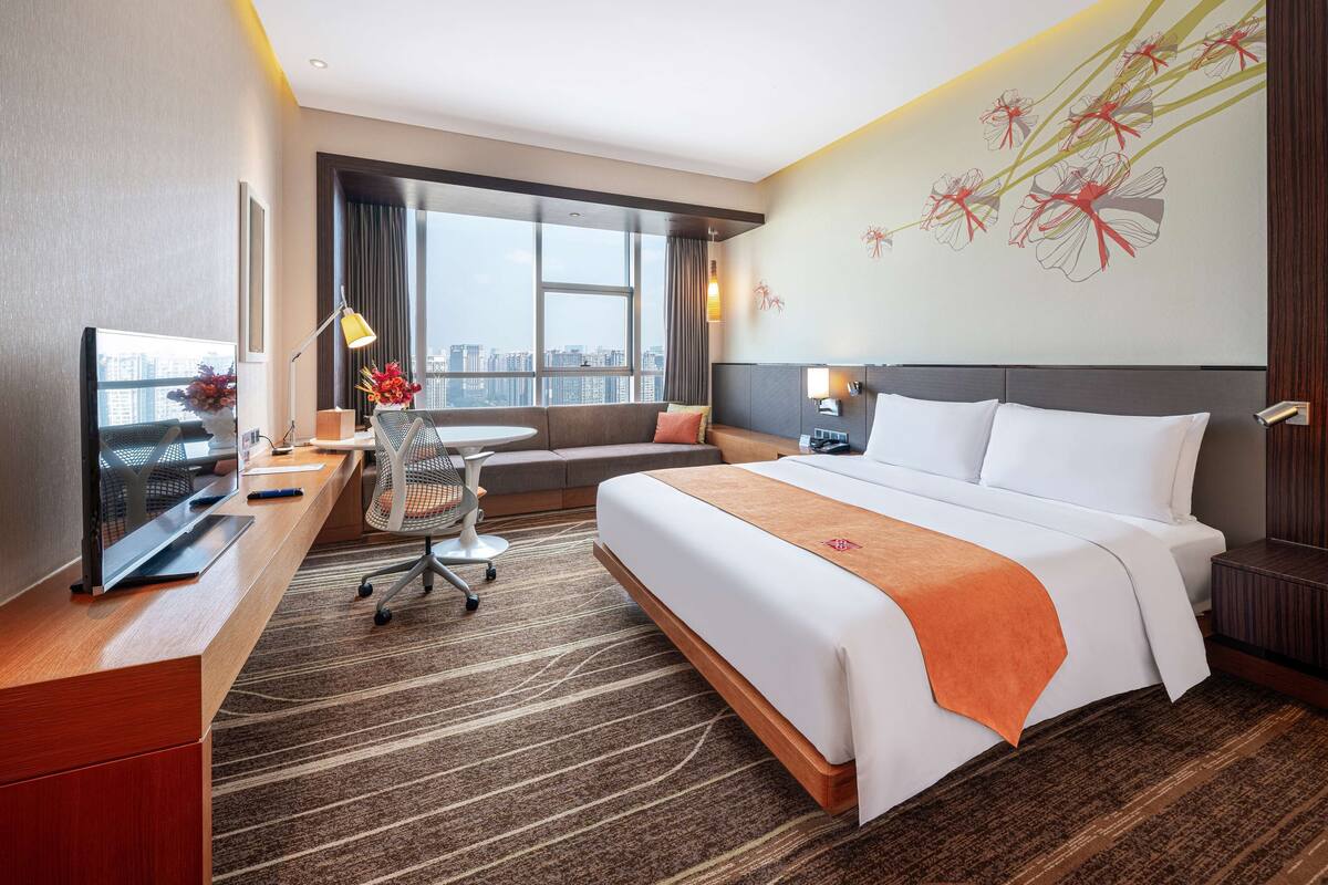A hotel room with a large bed, a desk with a computer, a chair, and a sofa. There is a large window with a view of the city.