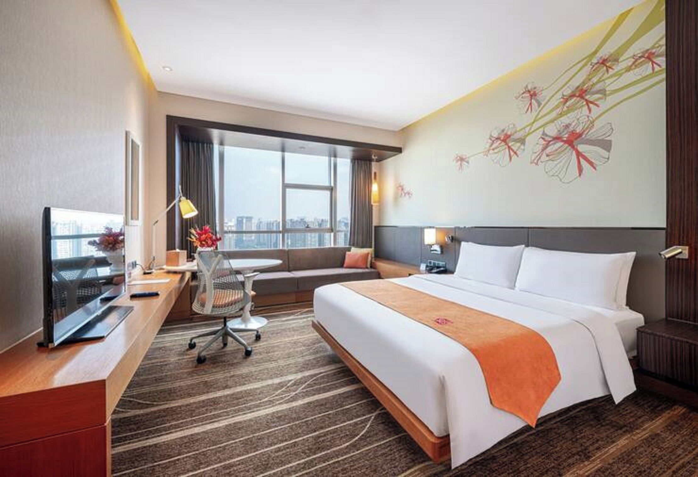 A hotel room with a large bed, a desk with a computer, a chair, a sofa, and a large window with a city view.