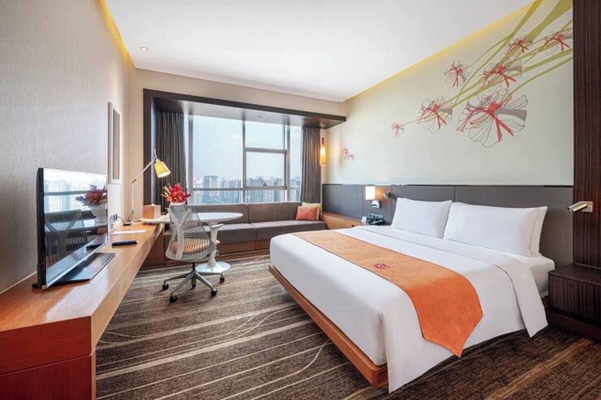A hotel room with a large bed, a desk with a computer, a chair, a sofa, and a large window with a city view.