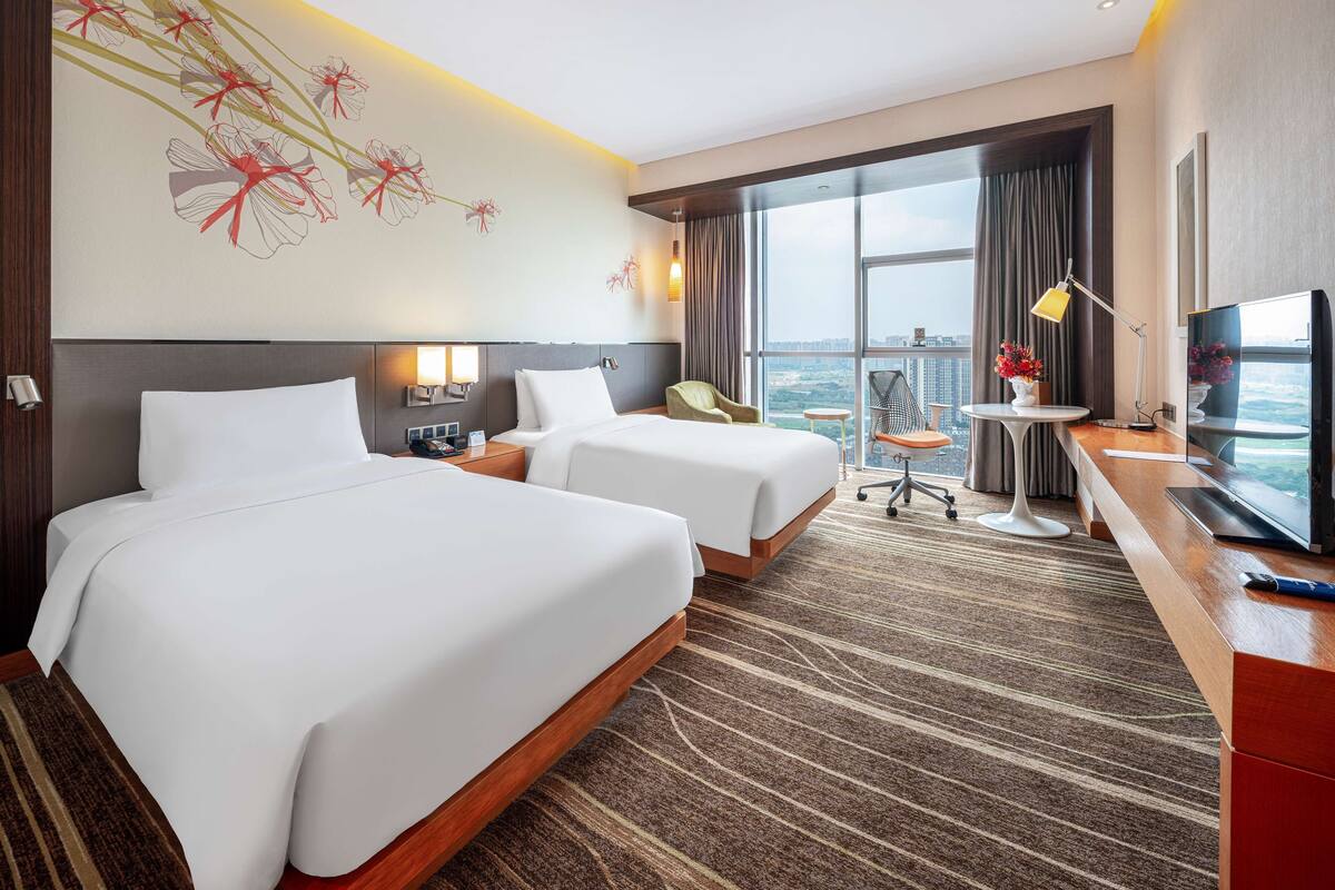 A hotel room with two beds, a desk with a TV, a chair, and a large window with a city view.