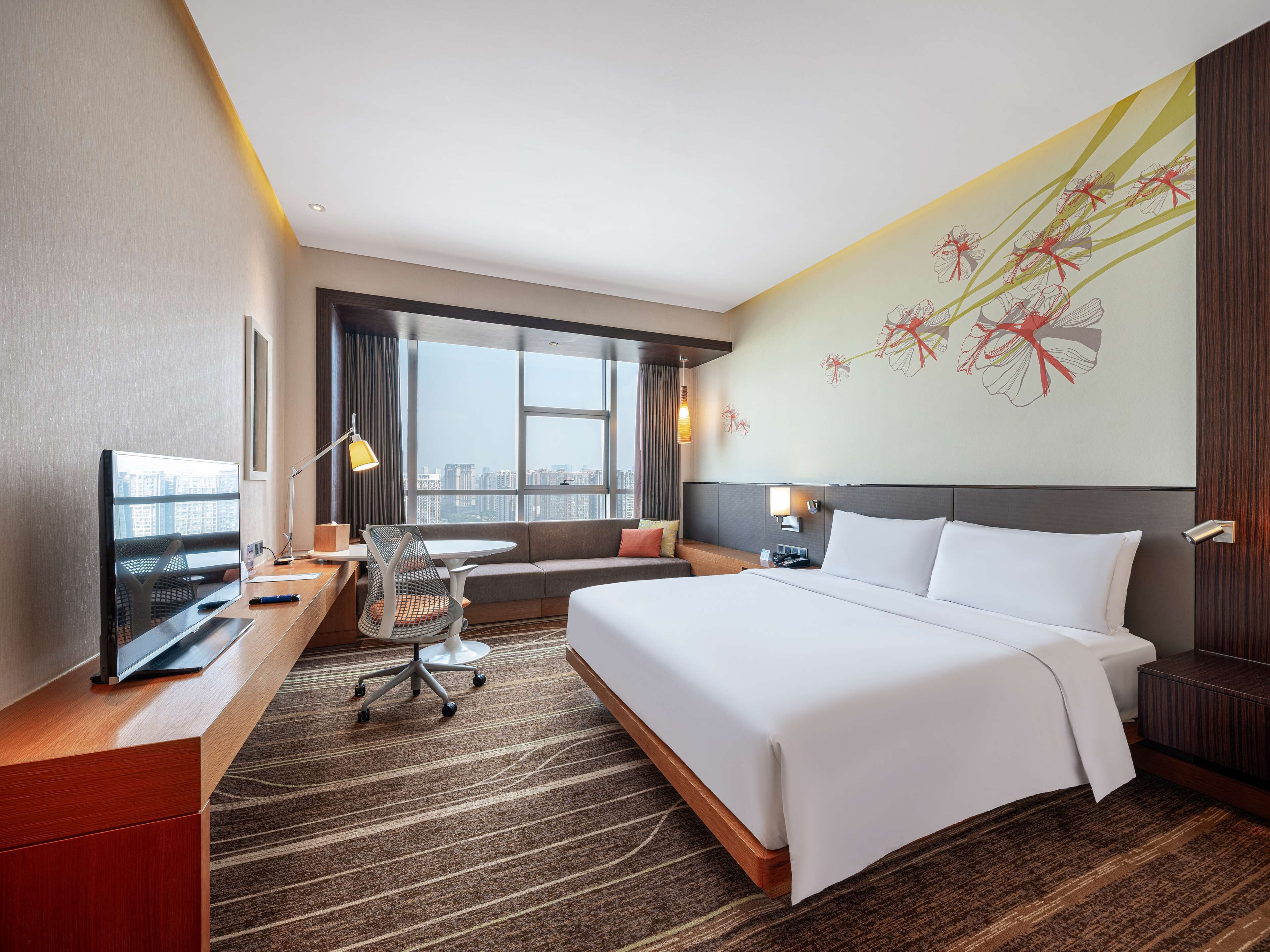 A hotel room with a large bed, a desk with a computer, a chair, a sofa, and a large window offering a city view.