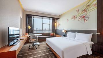 A hotel room with a large bed, a desk with a computer, a chair, a sofa, and a large window offering a city view.