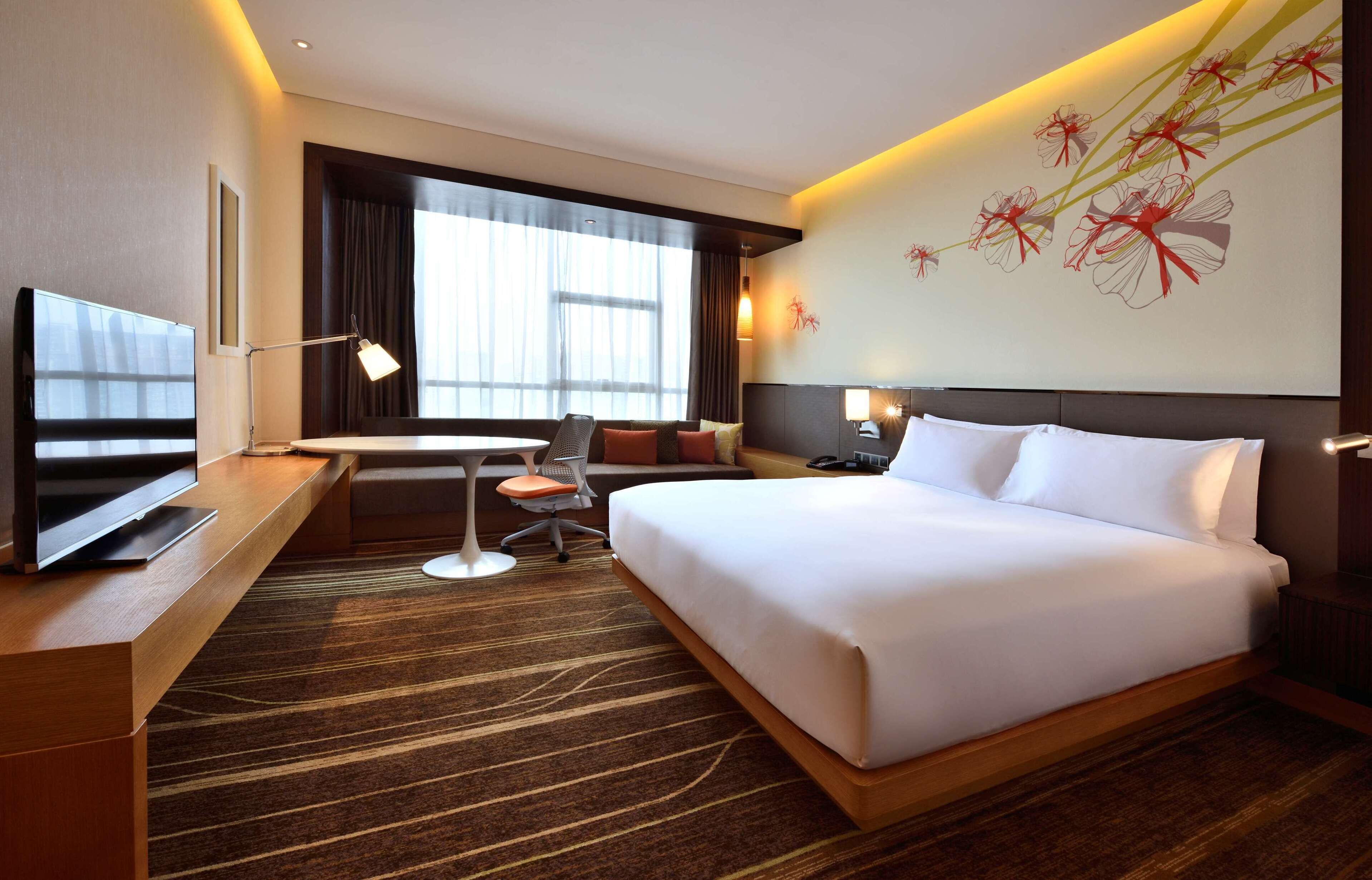 Habitación de hotel con una cama grande, un escritorio con televisor, una silla, una mesita y una ventana con cortinas.