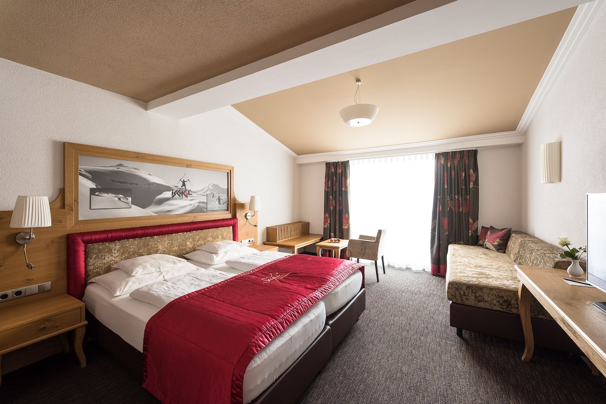 Quarto de hotel com cama, escrivaninha, cadeira, luminária, quadro e janela com cortinas.