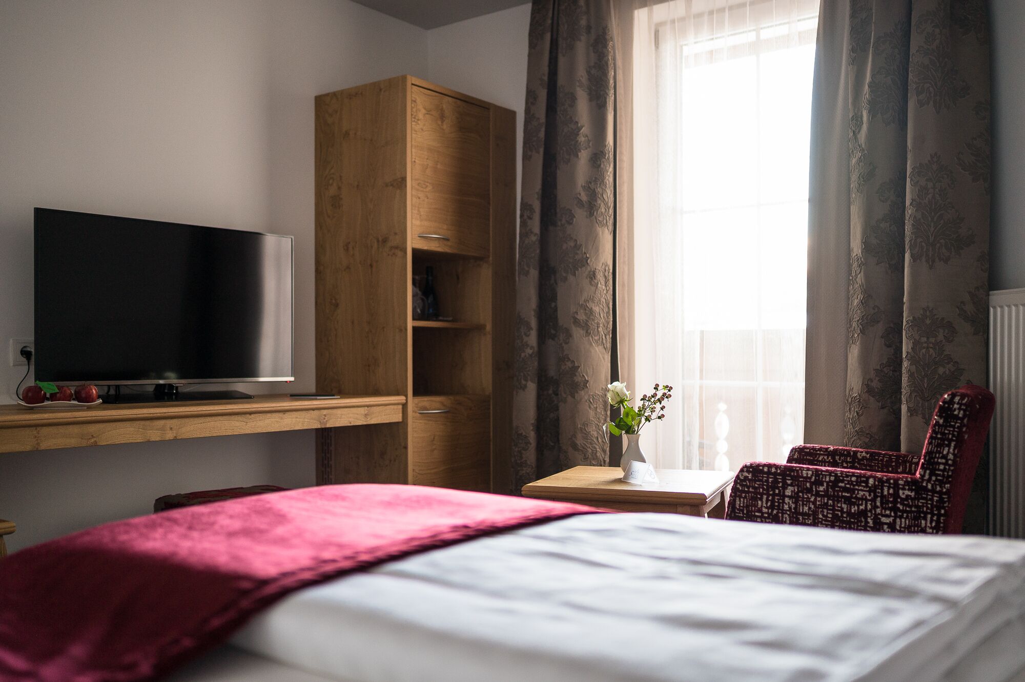 Habitación de hotel con cama, televisor en estante de madera, armario de madera, una mesita con un florero y una silla.