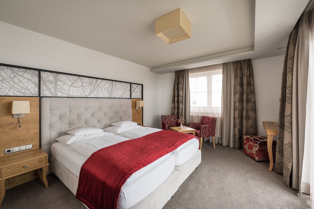 Quarto de hotel com uma cama grande, duas cadeiras, uma mesinha e uma janela com cortinas.