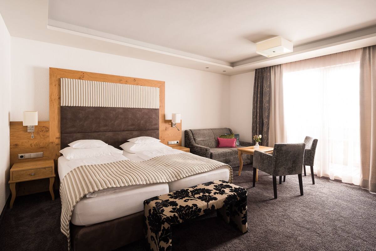 Quarto de hotel com cama, mesas de cabeceira, uma cadeira, uma mesinha e uma janela com cortinas.