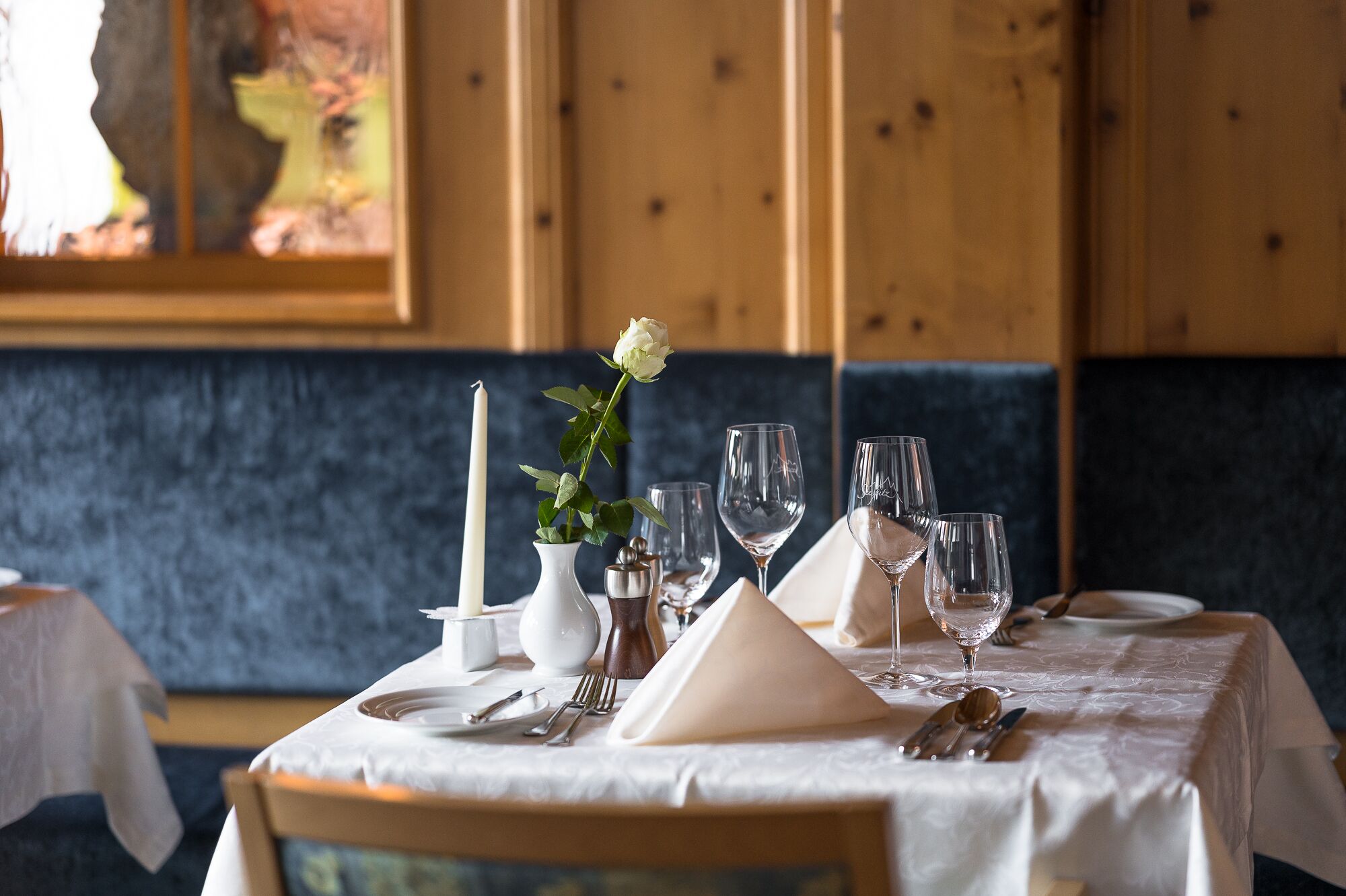 Una mesa de comedor dispuesta con mantel blanco, cubiertos y copas de vino, con una vela y un centro de mesa con una flor blanca.