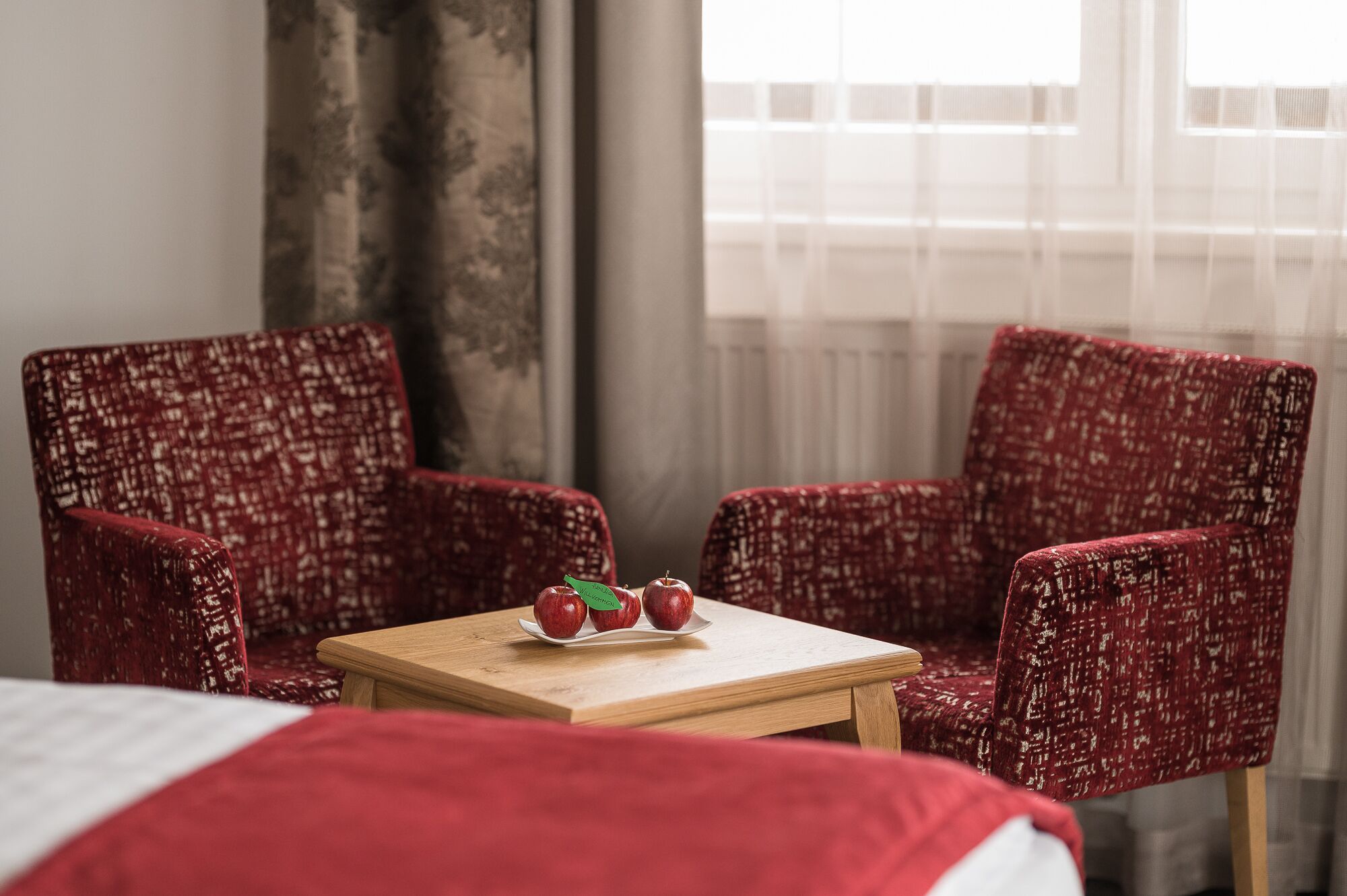 A hotel room with two red upholstered chairs, a wooden table with three apples, and a bed with a red blanket.