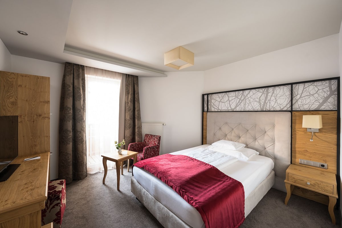 Quarto de hotel com cama, escrivaninha, cadeira, mesa de cabeceira e janela com cortinas.