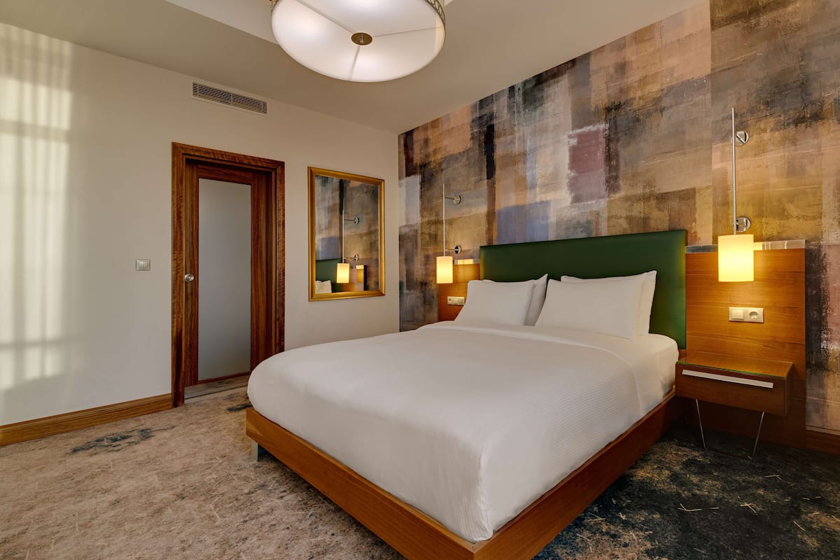 Habitación de hotel con una cama grande, un cabecero de madera, un espejo fijado a la pared y una mesita de noche con una lámpara.