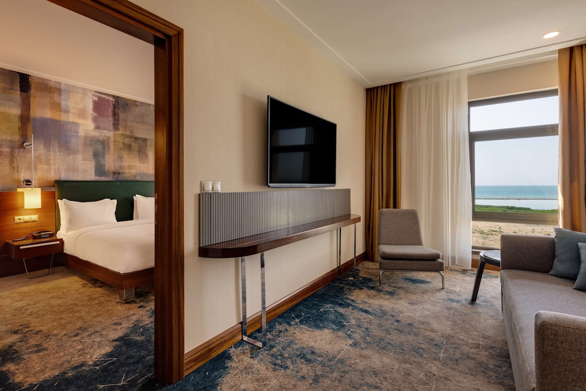 Habitación de hotel con cama, televisor montado en la pared, zona de estar con sillón y sofá, y vista al mar a través del ventanal.