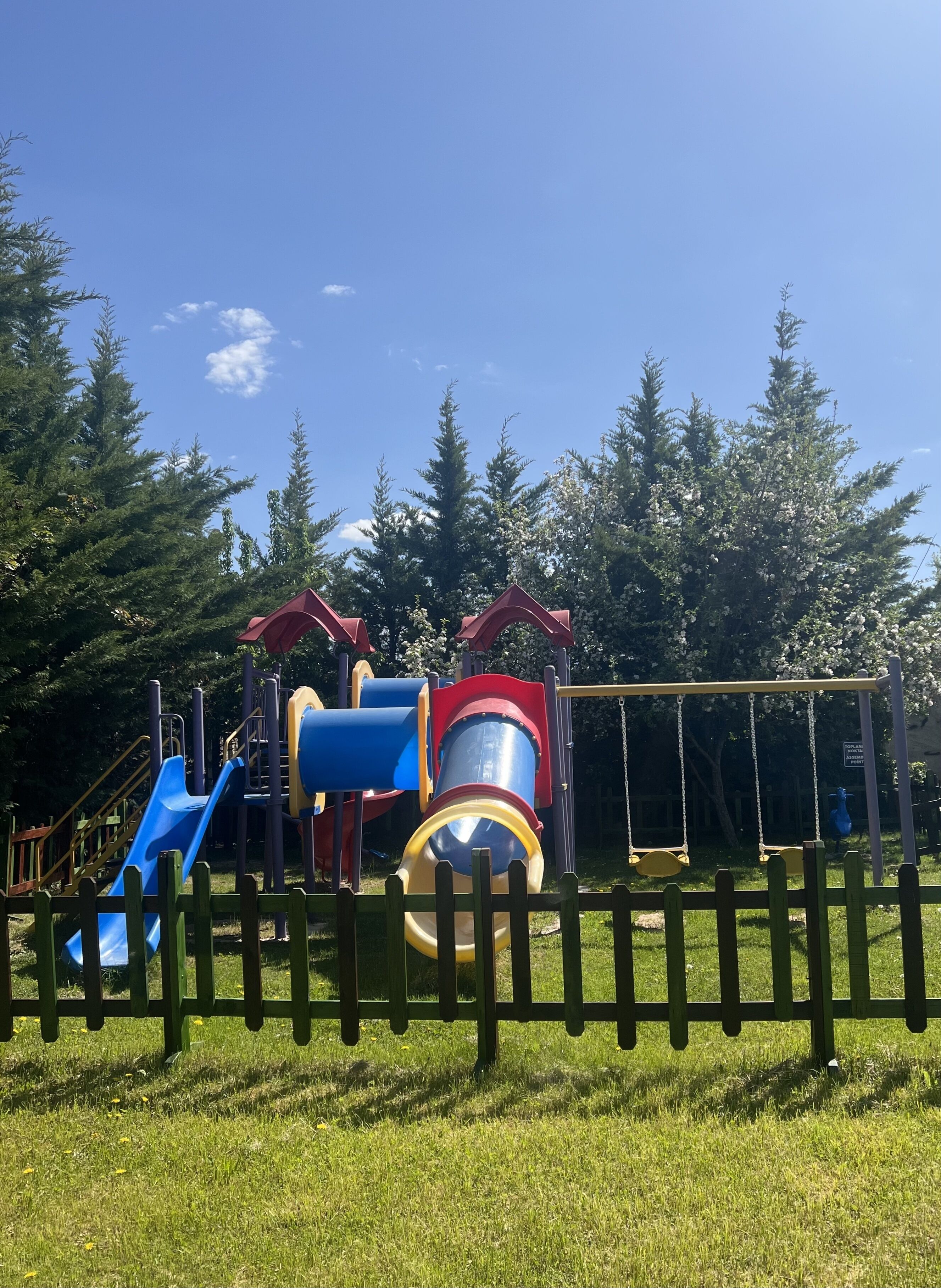 Un parque infantil con toboganes y columpios, rodeado por una valla de madera y árboles.