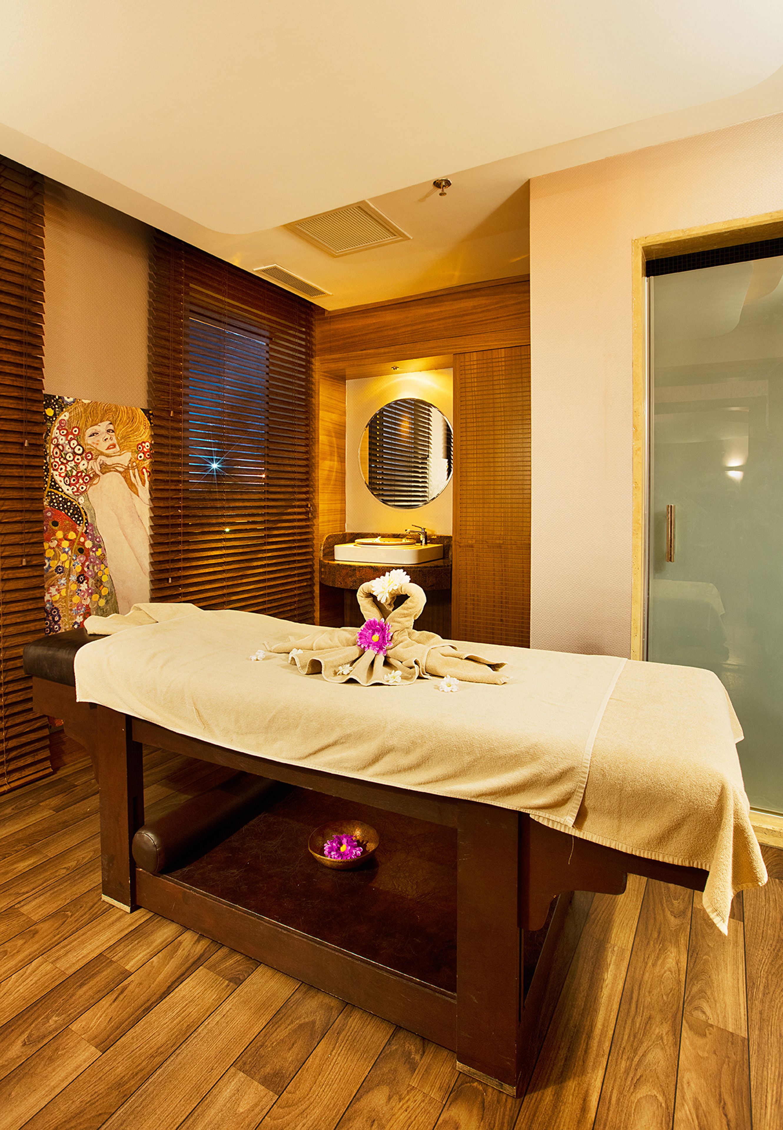 A massage table with towels and flowers, a wooden floor, a wall with a mirror, and a sliding glass door.