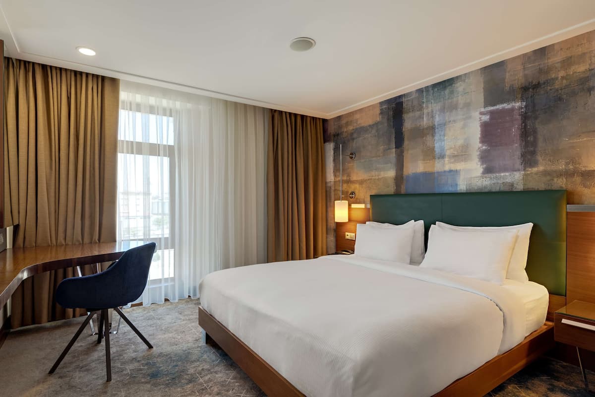 Habitación de hotel con una cama grande, un escritorio con una silla, un televisor y vistas de la ciudad a través de la ventana.