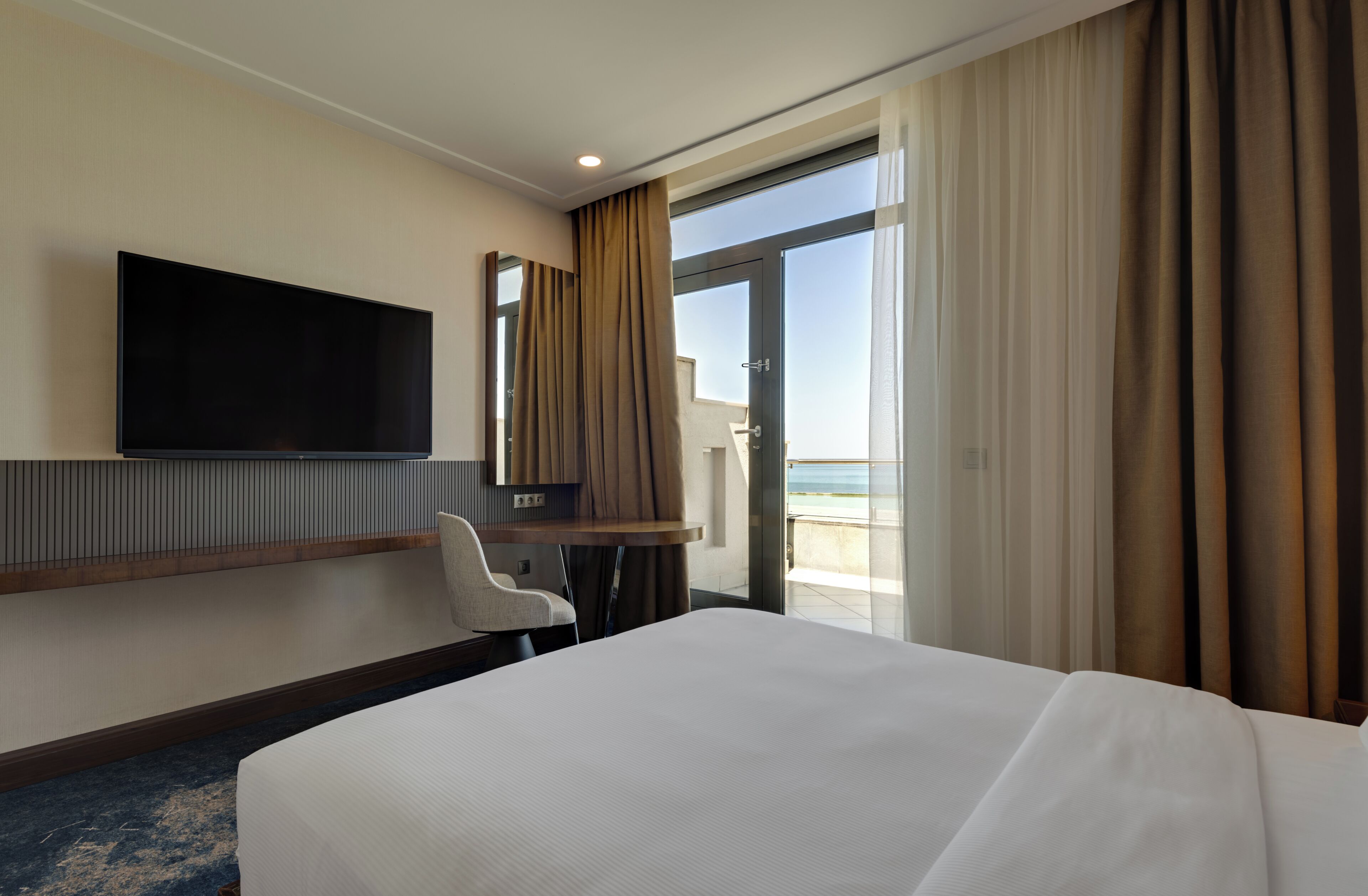 A hotel room with a large bed, a flat-screen TV mounted on the wall, a desk with a chair, and a balcony with curtains.