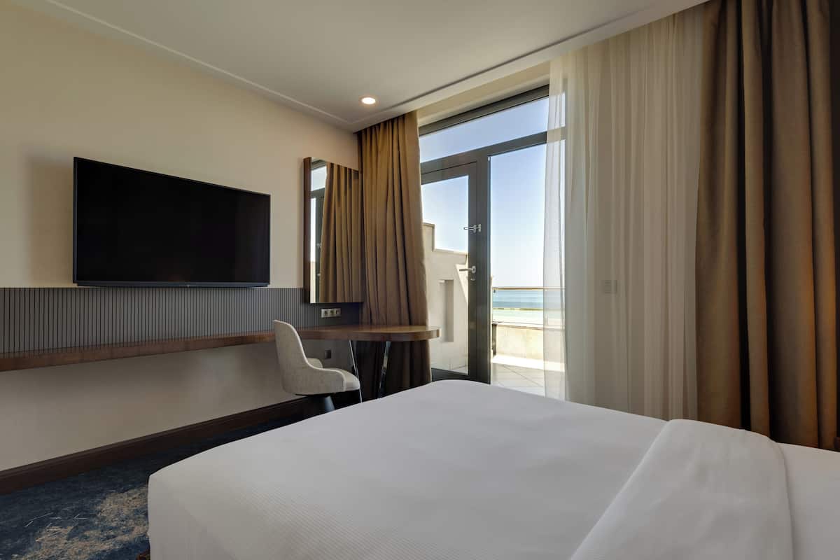 Habitación de hotel con una cama grande, un televisor de pantalla plana montado en la pared, un escritorio con silla y un balcón con cortinas.