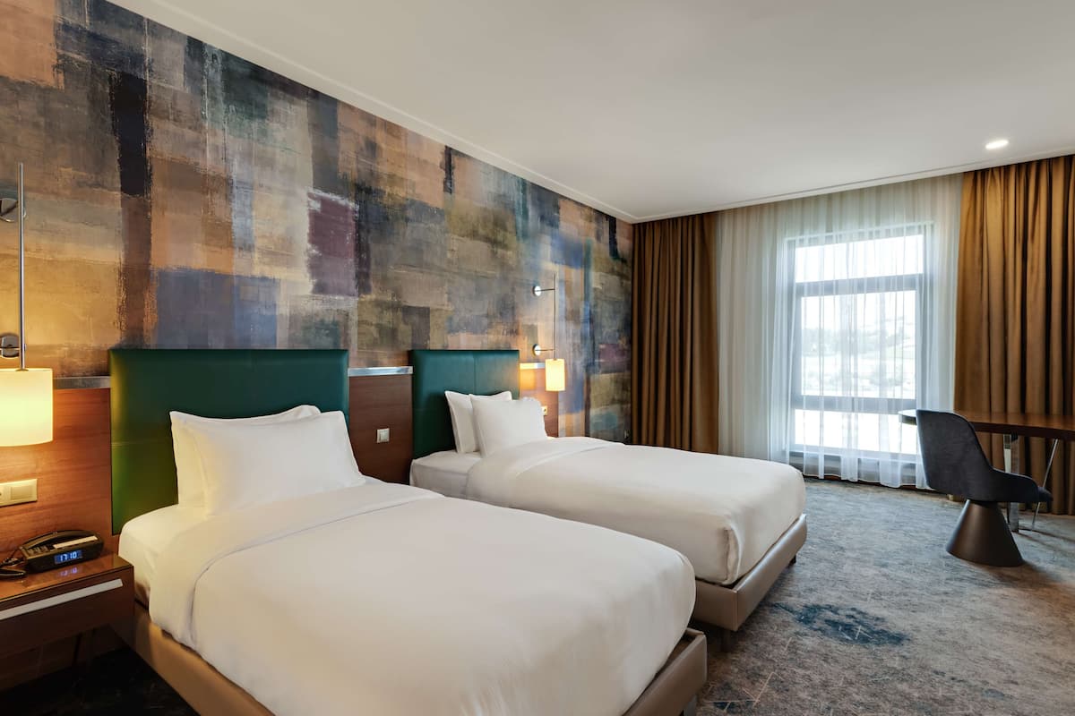 Habitación de hotel con dos camas, un escritorio, una silla y un ventanal con cortinas.