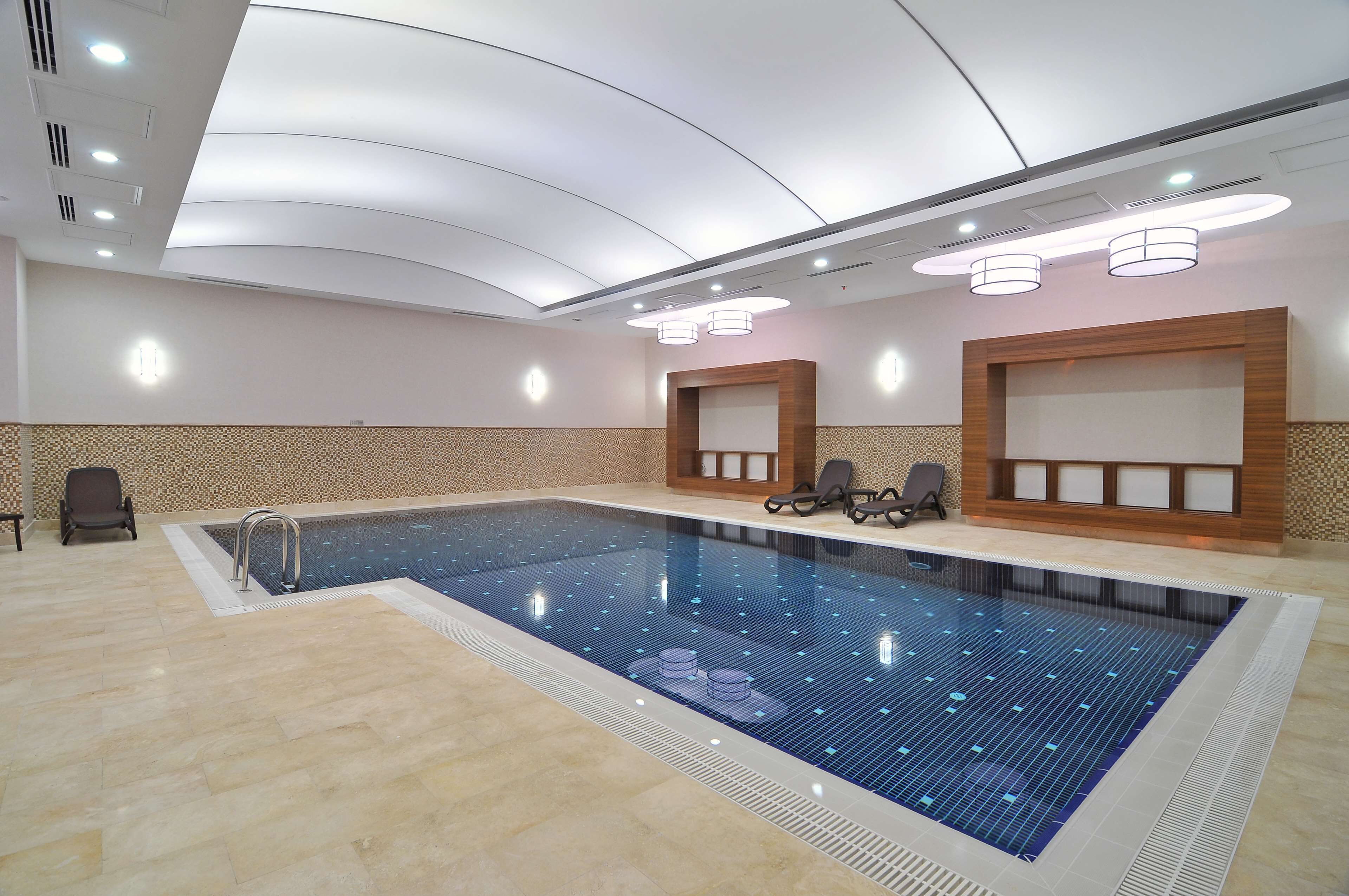 A large indoor swimming pool with a tiled floor, a seating area with chairs, and a ceiling with recessed lighting.
