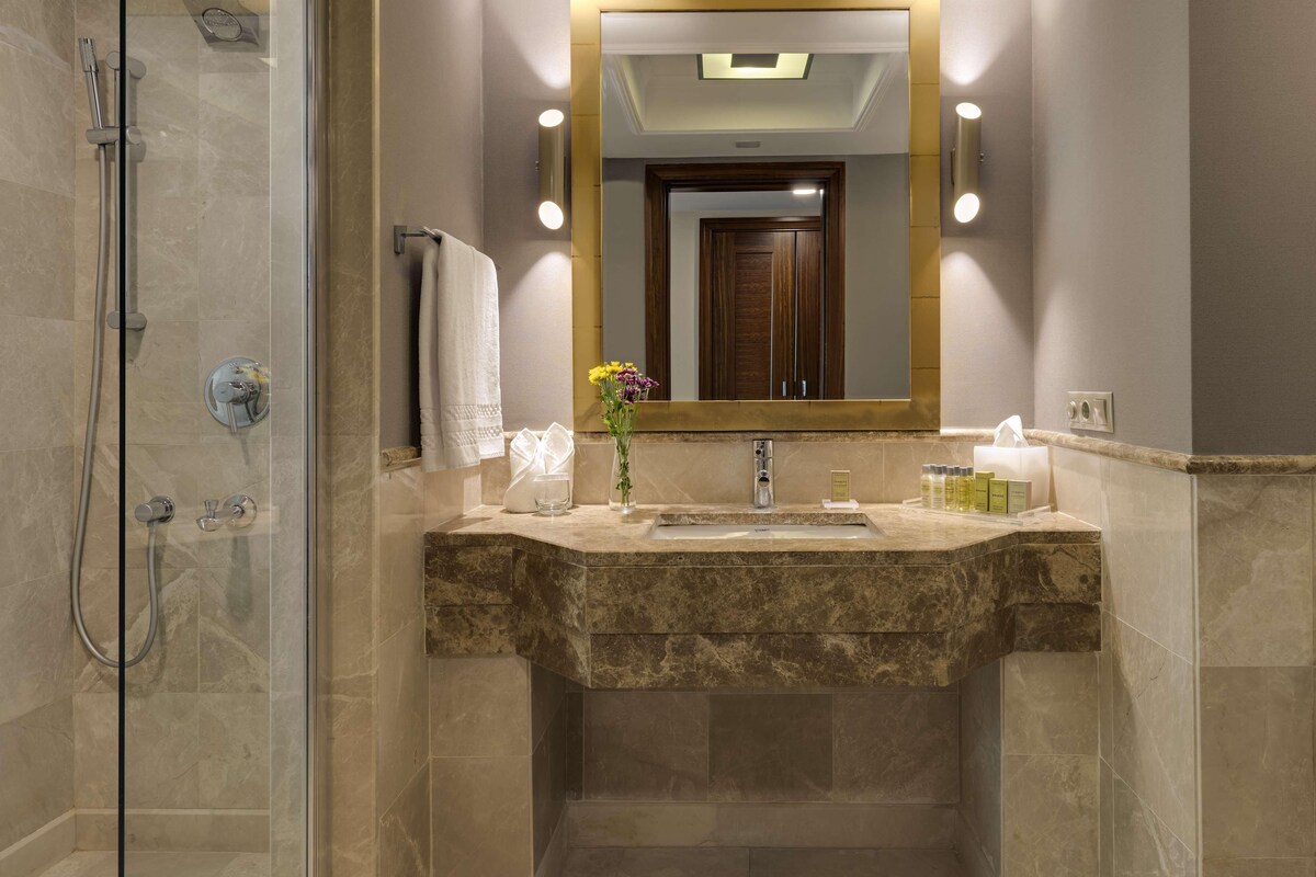 A bathroom with a glass-enclosed shower, a marble sink, and a mirror with wall sconces.