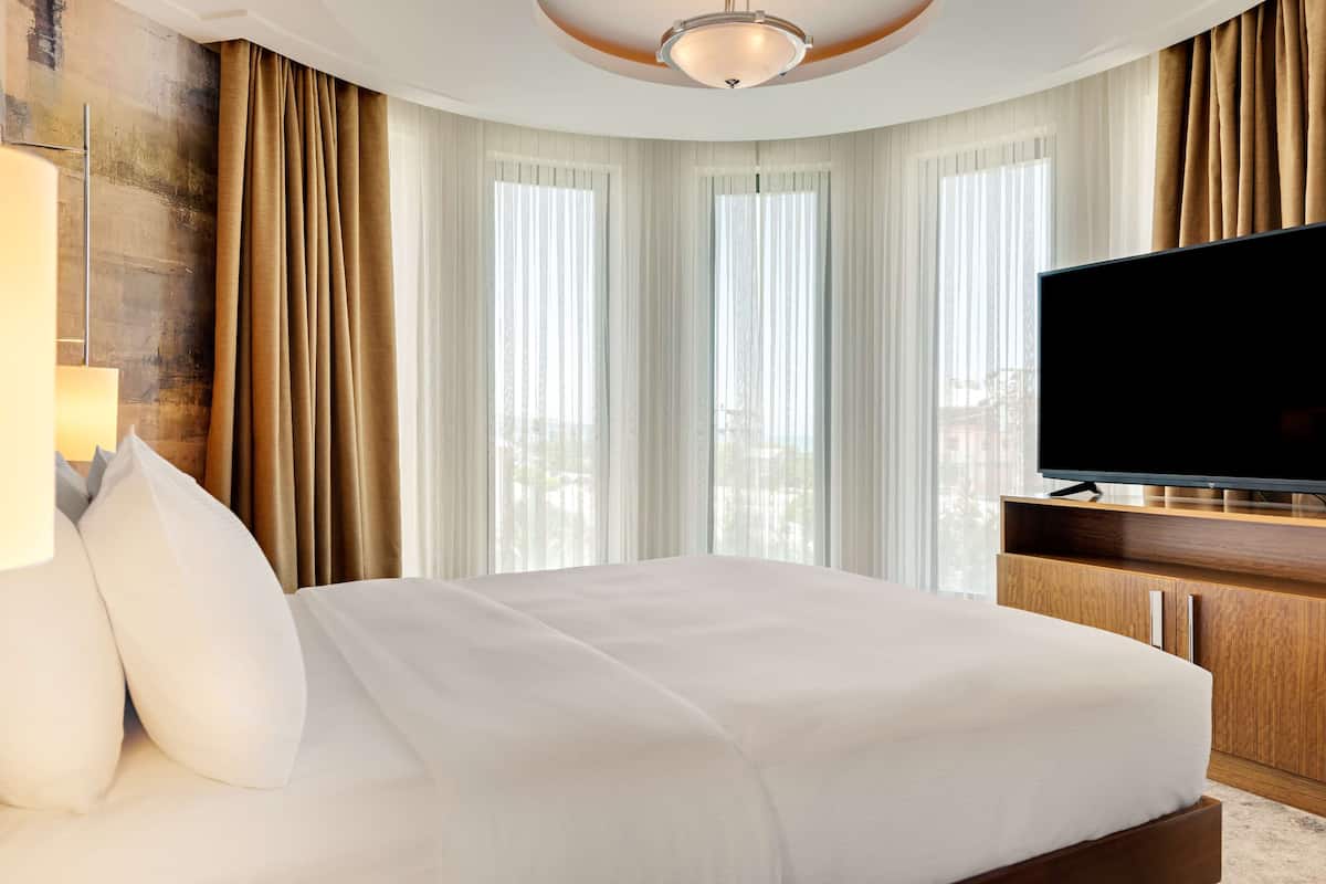 Habitación de hotel con una cama grande, un televisor de pantalla plana, muebles de madera y ventanas amplias con cortinas.