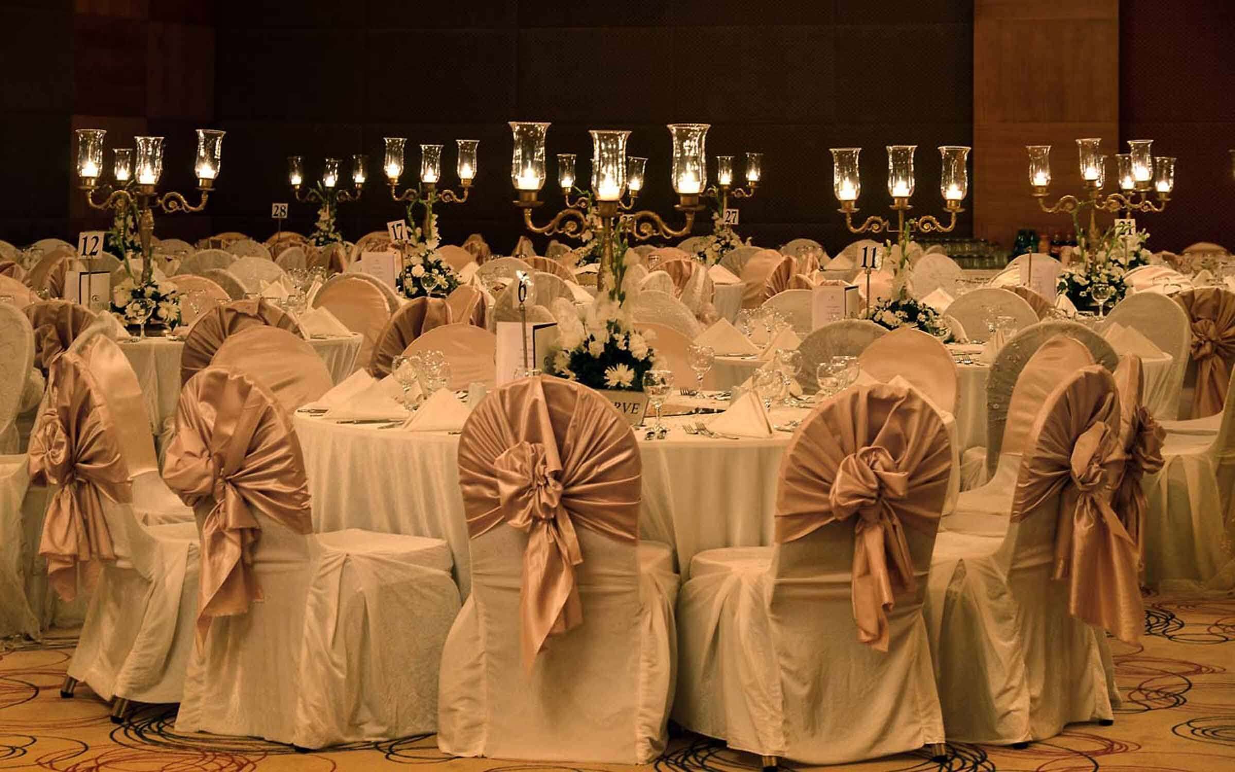 Un salón de banquetes con mesas redondas cubiertas con manteles blancos, cada una con un letrero con número. Las sillas están cubiertas de tela blanca y adornadas con cintas rosas.