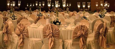 Un salón de banquetes con mesas redondas cubiertas con manteles blancos, cada una con un letrero con número. Las sillas están cubiertas de tela blanca y adornadas con cintas rosas.
