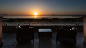 Un balcon avec deux fauteuils en osier et une petite table, d’où l’on peut admirer un coucher de soleil sur une étendue d’eau, avec des reliefs lointains à l’horizon.