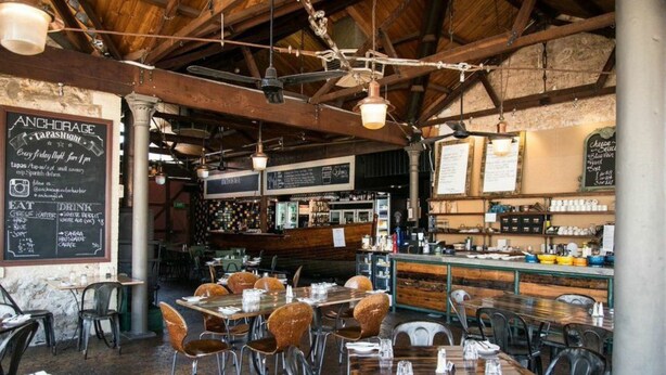 A restaurant interior with wooden beams, hanging lights, and tables with chairs. There are chalkboards with menus and a bar area in the background.