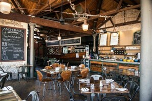 Interior de restaurante con vigas de madera, luces colgantes y mesas con sillas. Se observan pizarras con menús y una zona de bar al fondo.