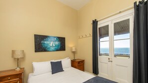A bedroom with a bed, two bedside tables, a painting, a window with curtains, and a view of the sea.